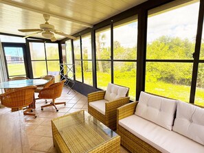 A sunlit room with a glass table, wicker furniture, and large windows offering a view of greenery. - Venice Villas Combo – Sleeps 5 | Poolside (Venice)