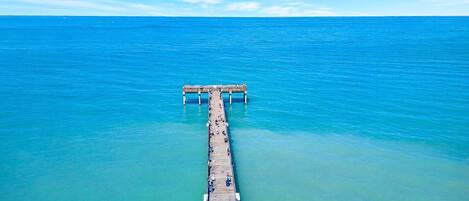 An aerial view of a pier extending into a calm sea with clear water, a wooden structure, and people walking along it.