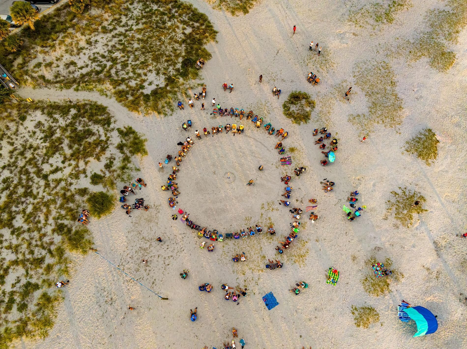 Eine Vogelperspektive eines Strandes, auf dem sich Menschen in einer kreisförmigen Anordnung versammelt haben, manche mit Strandausrüstung und Flaggen.