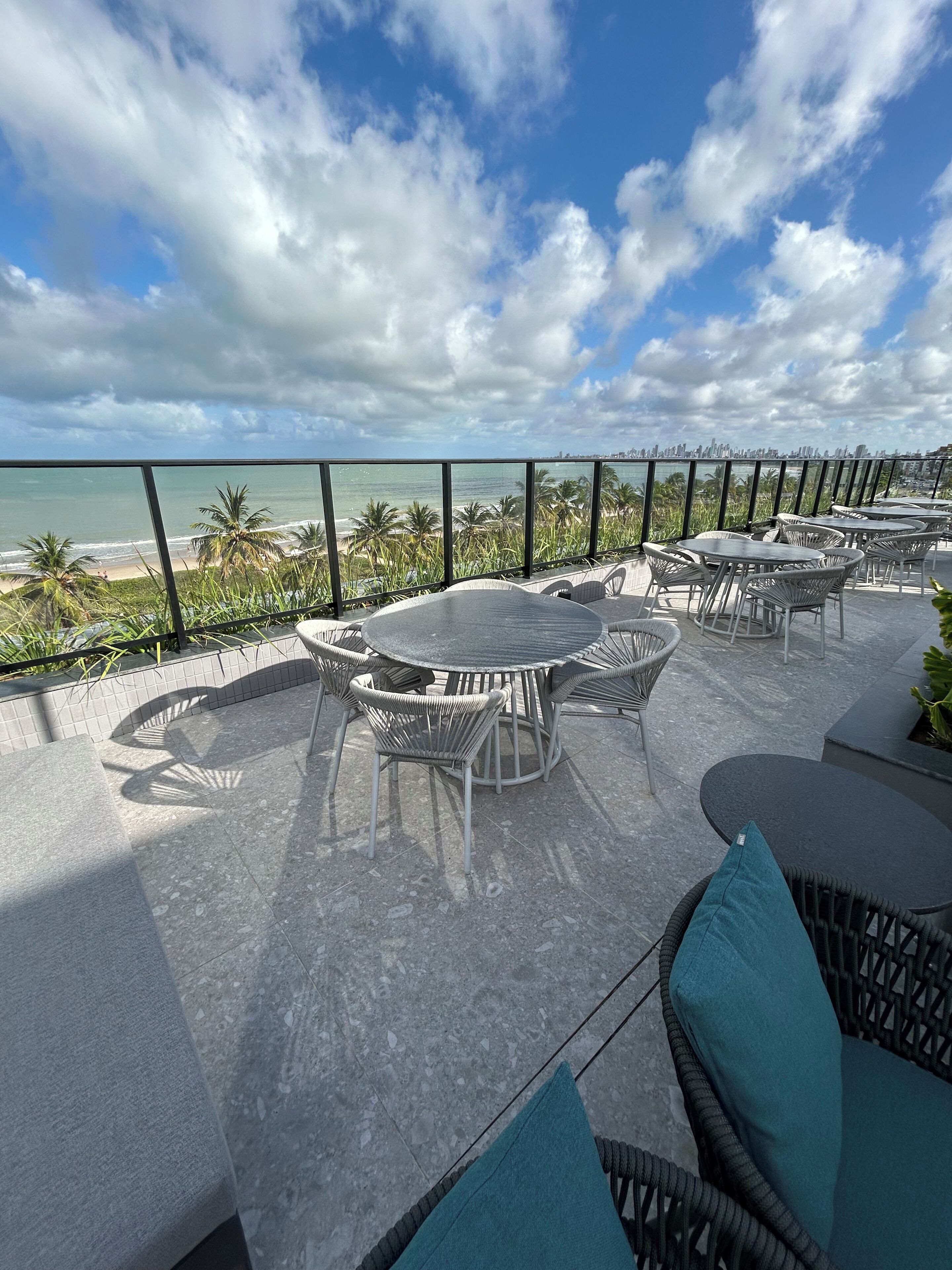 An outdoor patio with round tables and wicker chairs, overlooking a beach and city skyline.