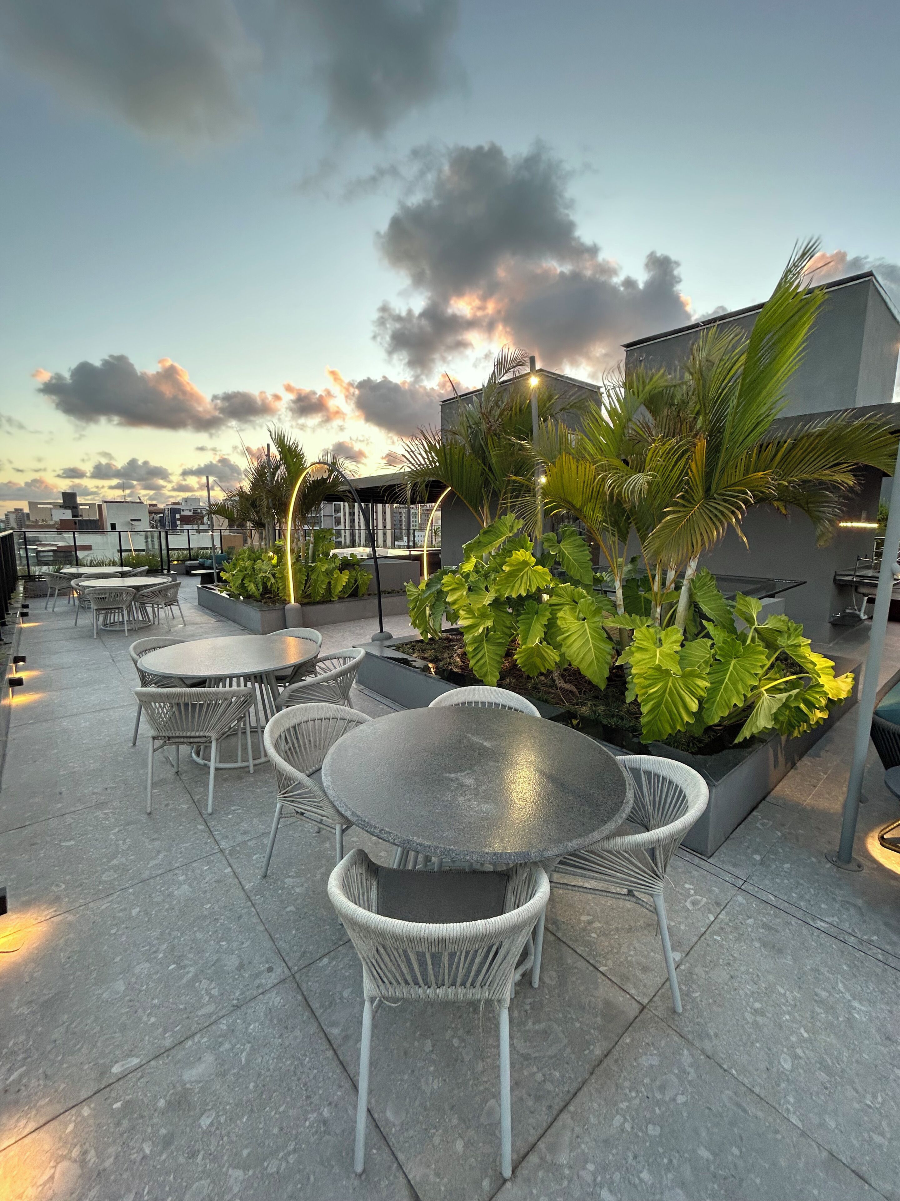 A rooftop terrace with outdoor seating, including round tables and wicker chairs, surrounded by lush greenery and palm trees.