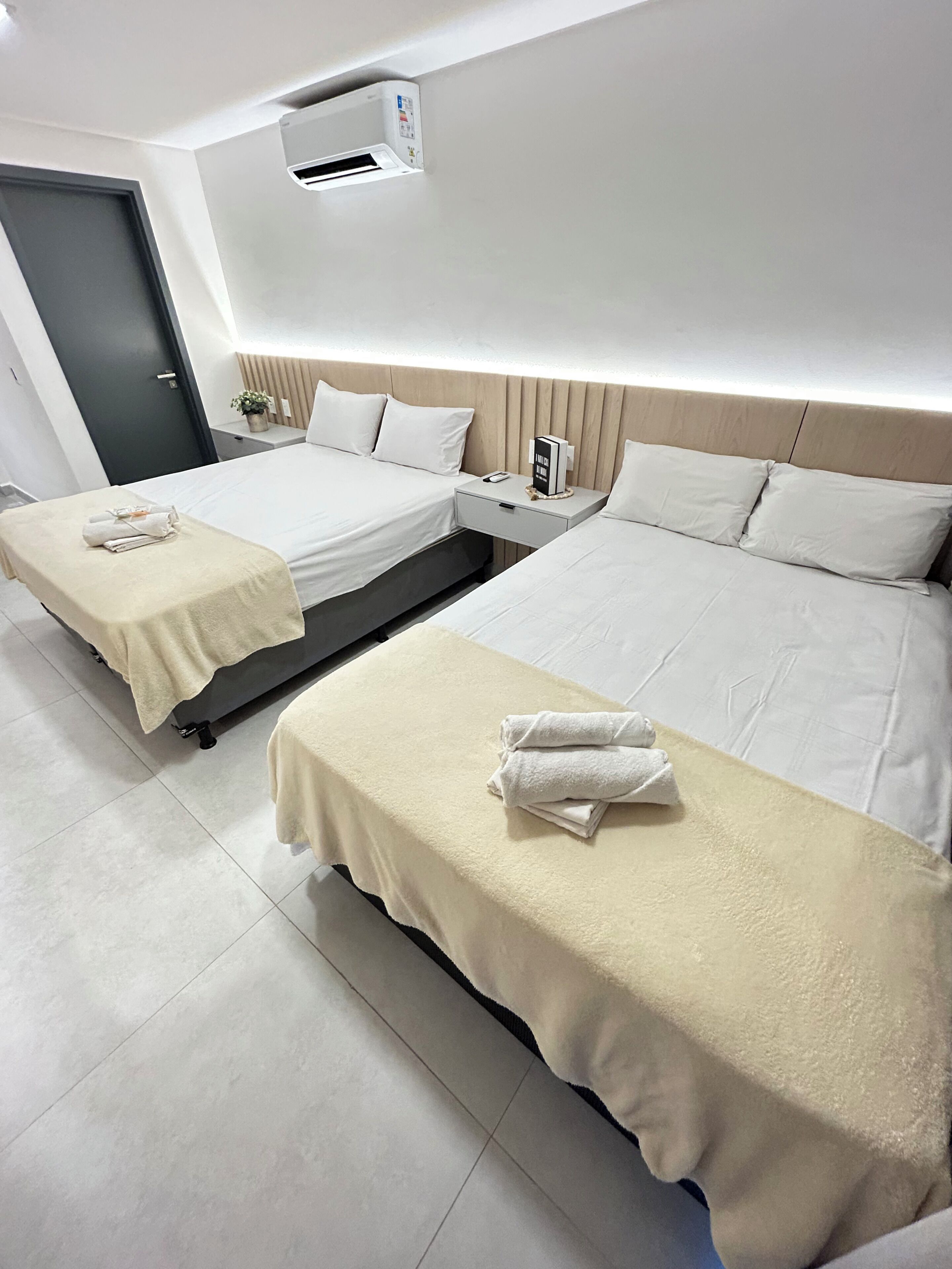 Hotellihuone, jossa on kaksi sänkyä, yöpöytä, jossa on puhelin ja kirja, seinälle asennettu ilmastointilaite ja ovi.