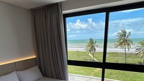 A hotel room with a large bed, a view of the beach, and a window with curtains. - Melhores Flats - Exclusividade vista mar (Cabedelo)