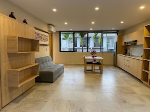Eine moderne Küche mit Holzausstattung, einem grauen Sofa, einem Esstisch mit Stühlen und einem großen Fenster mit Blick auf Palmen.