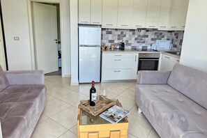 Eine moderne Küche mit weißen Schränken, einem Kühlschrank und einer Mikrowelle. Ein Holztisch mit einer Zeitschrift und einer Weinflasche steht bereit.