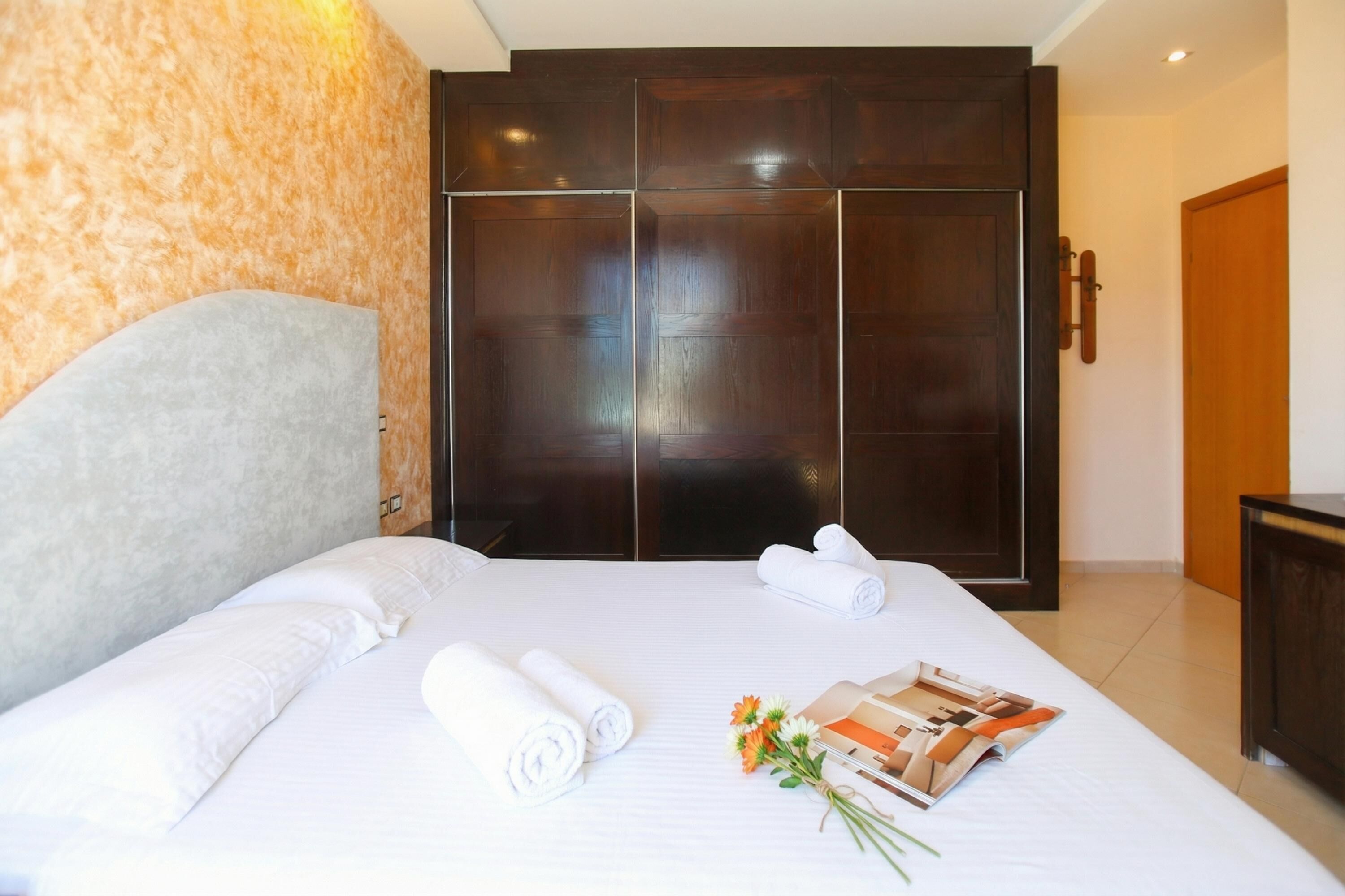 A hotel room with a large bed, a wardrobe, and a small table with a magazine and flowers.