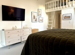 Un dormitorio con cama, televisor montado en la pared, cómoda, escalera y cuadros enmarcados.