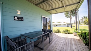 A covered outdoor seating area with a table and chairs, a sign reading 'The Beach House', and a view of a white building and palm trees in the background. - The Beach House - Tully Heads (Tully Heads)