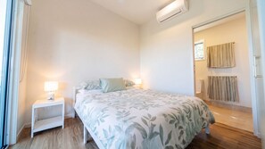 A bedroom with a bed, bedside lamps, a nightstand, a window, and an air conditioning unit. - The Beach House - Tully Heads (Tully Heads)