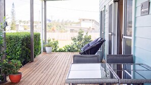 A wooden deck with a table and chairs, a potted plant, and a sliding glass door leading to a room. - The Beach House - Tully Heads (Tully Heads)