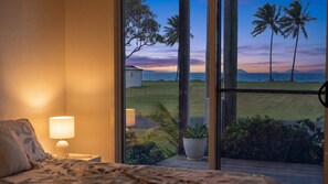 A bedroom with a large window overlooking a grassy area with palm trees and a view of the sea at sunset.