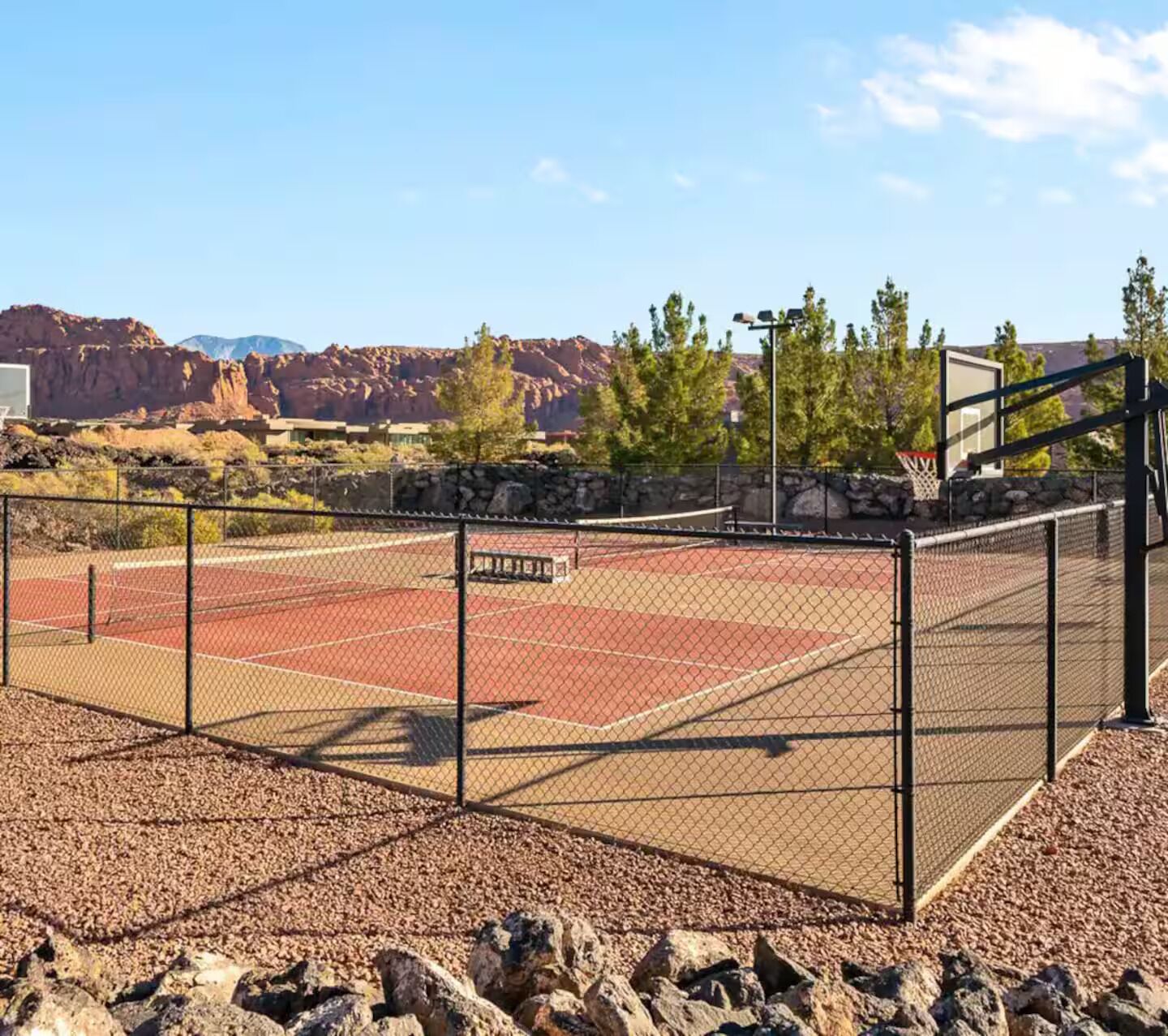 Ein Basketballplatz im Freien mit einem Drahtzaun, umgeben von Steinen und Bäumen, mit Bergen im Hintergrund.