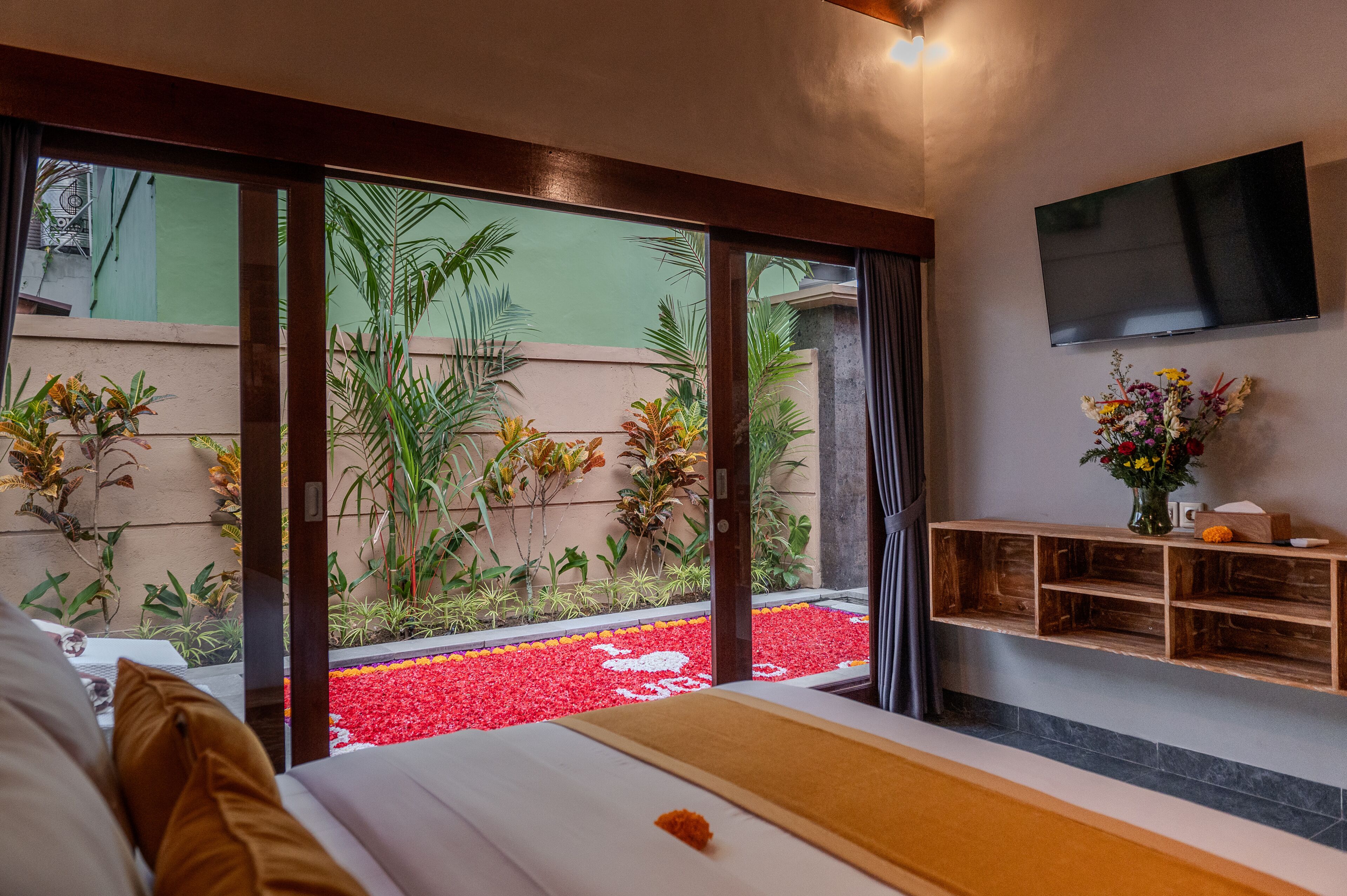 Un dormitorio con cama, mesa de masaje, televisor y vistas a un jardín con plantas y un parterre rojo.