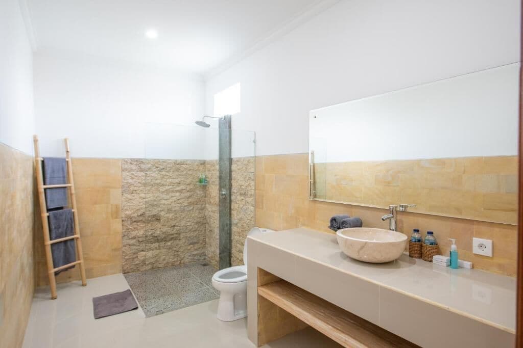 Een badkamer met een stenen douche, toilet, een wastafel met grote spiegel en een houten plank.
