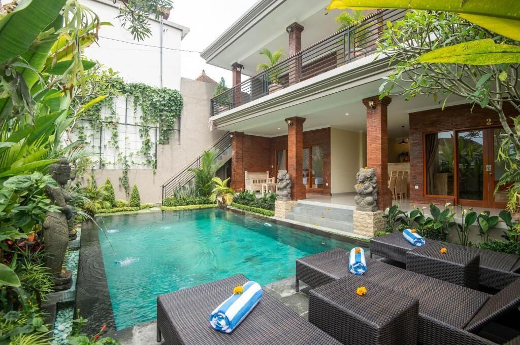 Una piscina rodeada de vegetación, un porche cubierto y un edificio de dos pisos.