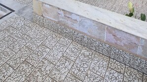 Um espaço ao ar livre coberto, com paredes de pedra, duas cadeiras de madeira, um banco e vasos com plantas.
