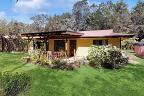 A single-story house with a covered porch, a well-maintained lawn, and a garden with various plants and flowers. - Two charming homes on one lot - nature walks, meditation spots and gardens (Volcano)