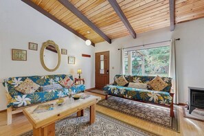 A living room with a sofa, a coffee table, a fireplace, and a large window with a view of greenery. - Two charming homes on one lot - nature walks, meditation spots and gardens (Volcano)