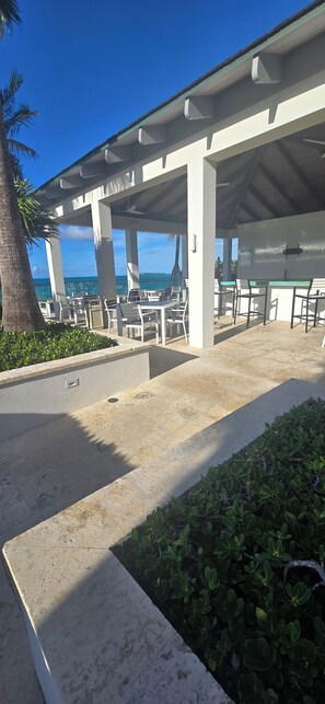 A covered outdoor seating area with tables and chairs, a palm tree, and a view of the ocean. - Paradise at one cable beach (Nassau)
