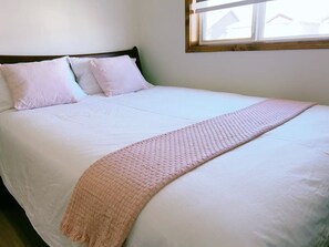 A bed with white bedding and a pink knitted throw, a wooden headboard, and a window with a view of houses. - Modern 3BR Panorama Home | Garage | Near YYC (Calgary)