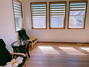 A room with three large windows, a round table with a chair, and a sofa. - Modern 3BR Panorama Home | Garage | Near YYC (Calgary)