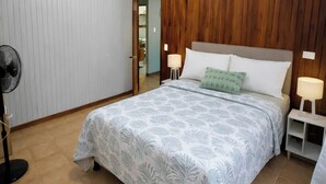A bedroom with a wooden paneled wall, a bed with a patterned bedspread, two bedside lamps, and a standing fan.