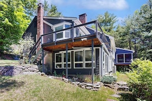 A modern house with a large deck, a chimney, and a stone retaining wall. - Stunning home with amazing views, a private pool, serenity & lake access (Meredith)