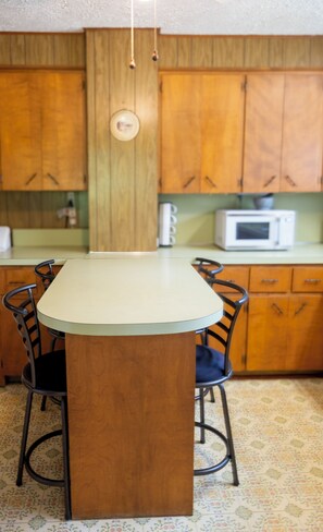 Een keuken met houten kastjes, een witte magnetron en een centraal kookeiland met stoelen.