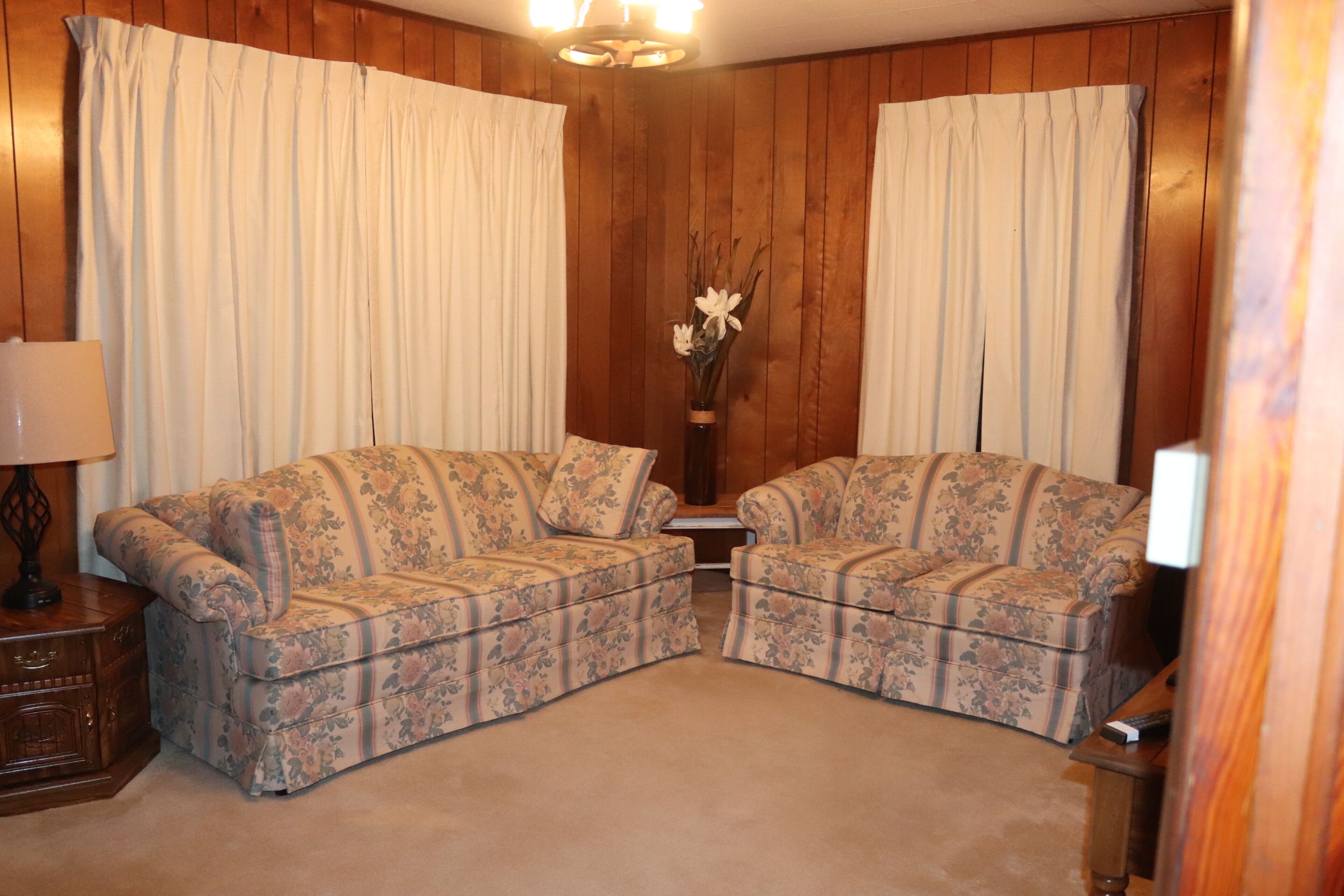 Una habitación con dos sofás de estampado floral, un aparador de madera, un jarrón con flores y una lámpara en el techo.