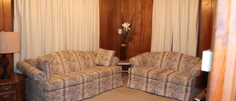 Una habitación con dos sofás de estampado floral, un aparador de madera, un jarrón con flores y una lámpara en el techo.