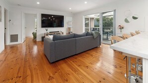 A modern living room with a grey sofa, wooden flooring, a flat-screen TV, and a dining area with a white table and chairs. - Foreshore Hideaway - Capel Sound (Capel Sound)