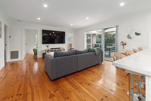 Ein modernes Wohnzimmer mit grauem Sofa, Holzboden, Flachbildfernseher und einem Essbereich mit weißem Tisch und Stühlen.