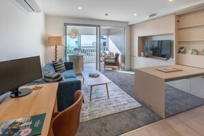 A modern living room with a sofa, coffee table, and a desk with a computer. There is a balcony with a view. - Waves & Wellness |Spa Pool, Sauna + Parking (Christchurch)