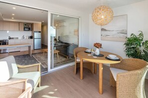 A modern living space with a dining area, kitchen, and sliding glass doors leading to a balcony. - Waves & Wellness |Spa Pool, Sauna + Parking (Christchurch)