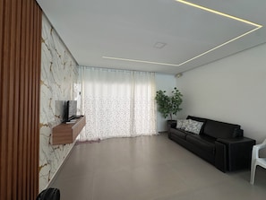 A modern living room with a flat-screen TV, a dark sofa, a potted plant, and a window with sheer curtains. - Beach house in Barra de Jacuípe, 5 bedrooms, ideal for families, near Praia do Forte. (Camaçari)