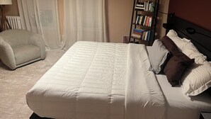 Una cama bien hecha con ropa blanca, una estantería con libros, una silla y una lámpara.