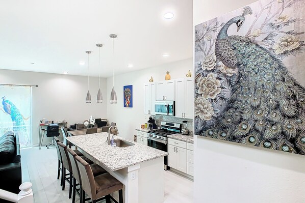 A modern kitchen with white cabinets, a central island, and a peacock painting on the wall. - 4 Bed Villa-sleeps 8-pool-gym-15 min to Disney (Kissimmee)