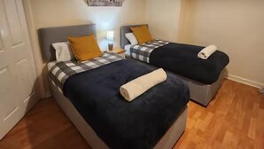 Two single beds with dark blue bedding and white pillows, a wooden floor, and a white door. - Comfy 3-bed House - Sleeps 6 & Free Parking (Tilbury)