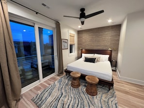 Ein Schlafzimmer mit Bett, Deckenventilator, Nachttisch und Fenster mit Vorhängen, mit Blick ins Freie.