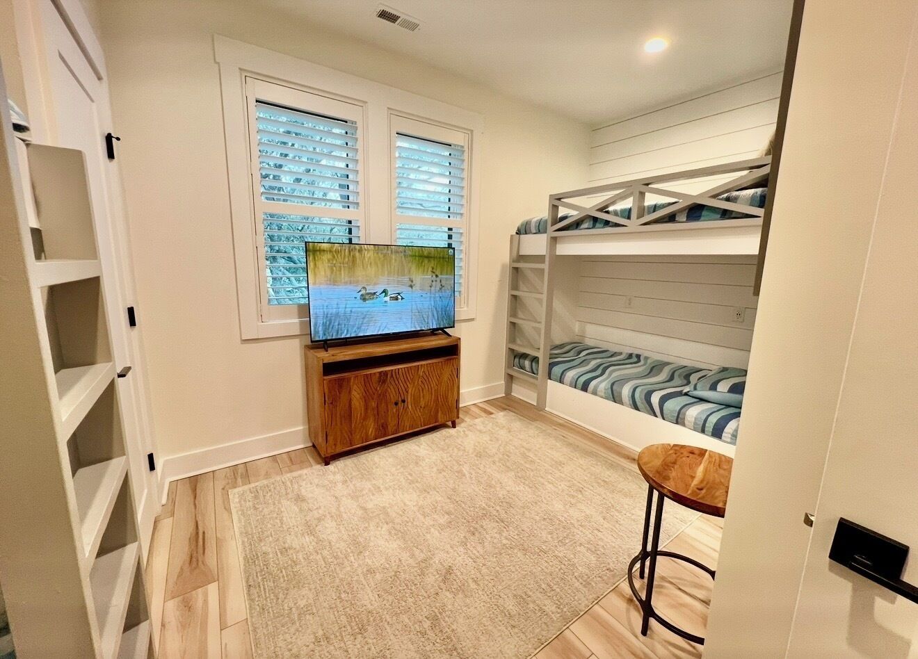 Une chambre avec un lit superposé, un téléviseur posé sur un meuble en bois, une petite table et une fenêtre avec des stores.