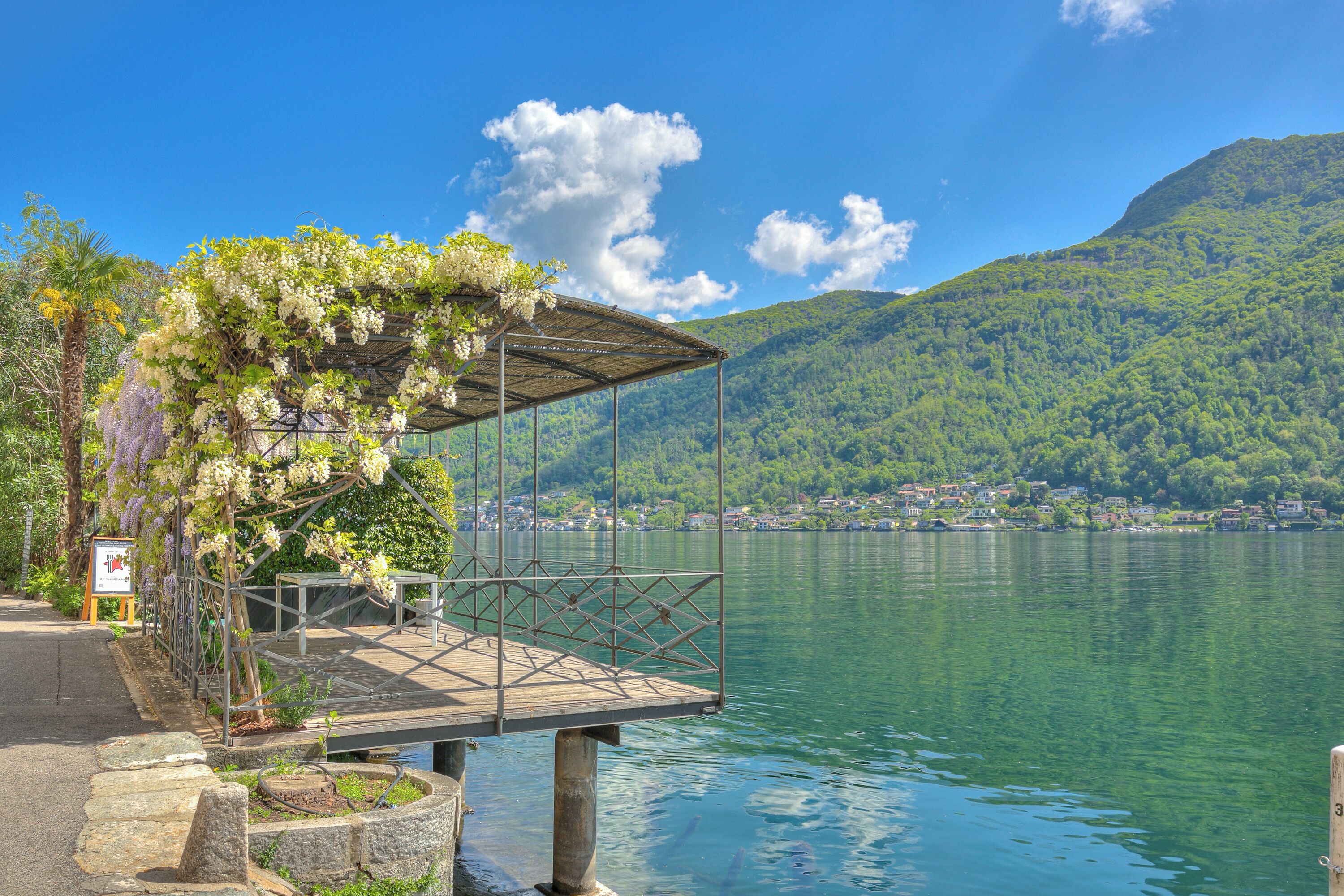 Gazebo sul lago con tetto in metallo, circondato da vegetazione lussureggiante e una vista limpida sul lago e sulle montagne in lontananza.