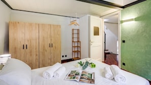 Un letto rifatto con cura, asciugamani arrotolati, una rivista e una piccola pianta. Si vede un armadio in legno e una scala appoggiata al muro.