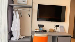 Ett hotellrum med en säng, ett skrivbord med en stol, en tv, en termostat och en papperskorg.