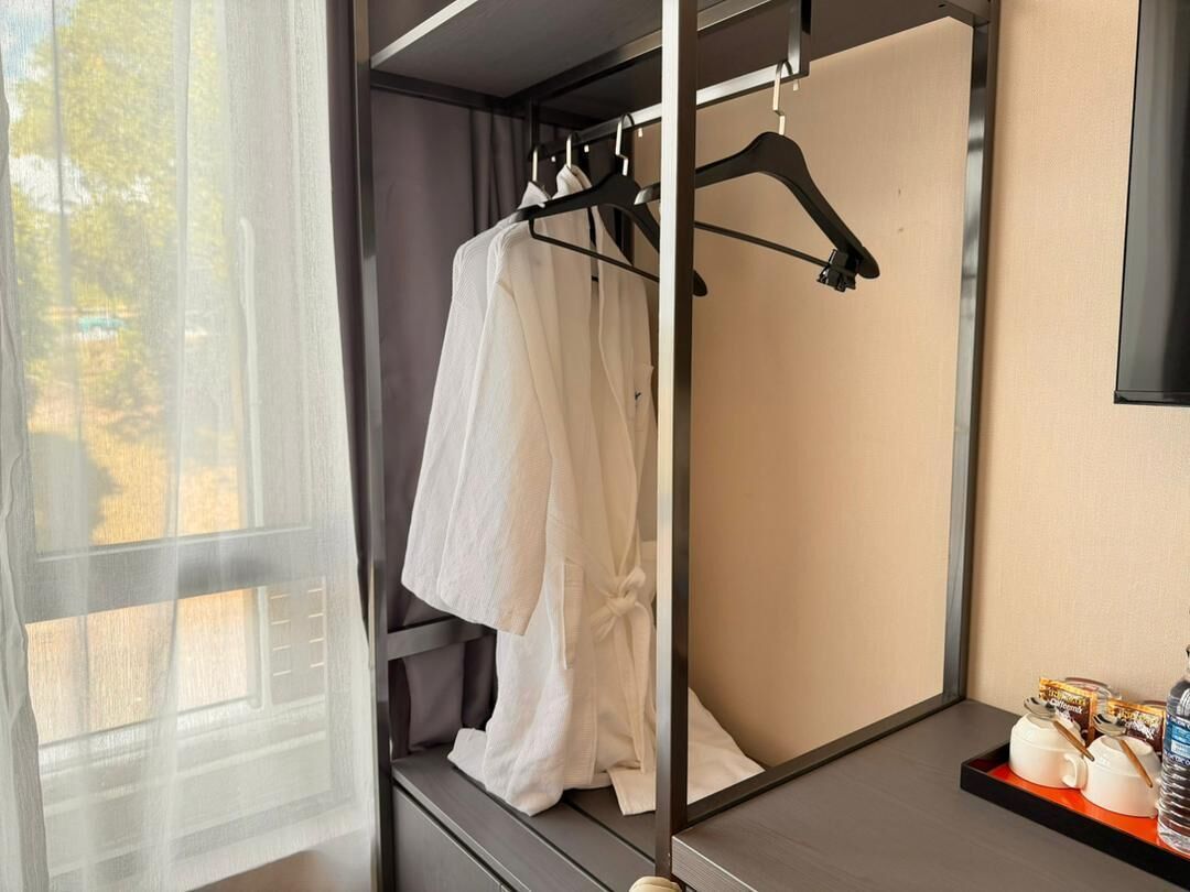 A hotel room with a bed, a window with sheer curtains, a robe hanging on a rack, and a tray with toiletries.