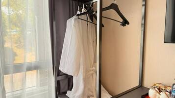 A hotel room with a bed, a window with sheer curtains, a robe hanging on a rack, and a tray with toiletries.
