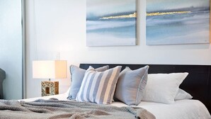 En pænt redt seng med stribede puder, et gråt tæppe og en hvid pude. Der er en lampe på et sengebord ved siden af sengen.