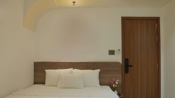 Quarto de hotel com cabeceira de madeira, roupa de cama branca, cama bem arrumada com toalhas, mesinha de cabeceira com um vaso de flores e porta de madeira.