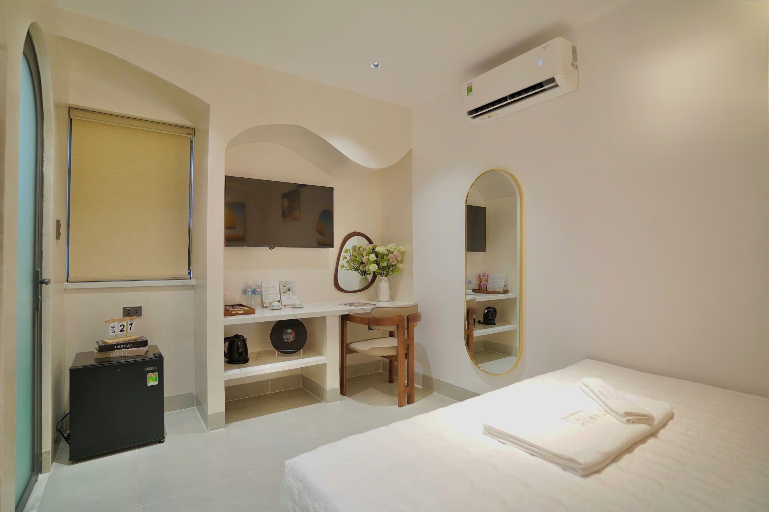 A hotel room with a bed, a desk with a chair, a mirror, a small refrigerator, and a shelf with toiletries.
