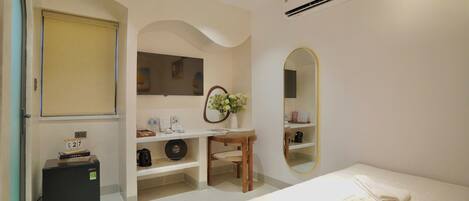 Habitación de hotel con cama, escritorio con silla, espejo, refrigerador pequeño y estante con artículos de aseo.