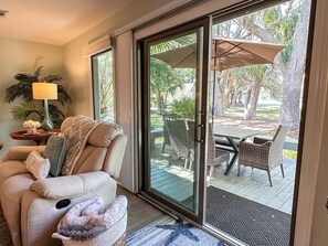 Ein Wohnzimmer mit einem beigefarbenen Sofa, einer Glastür zu einer Terrasse im Freien mit Korbmöbeln und einem Tisch.